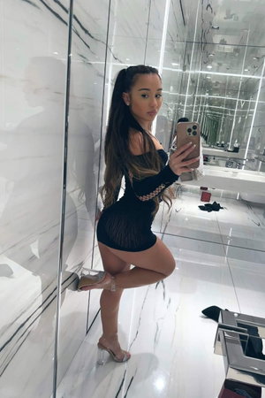 Asian woman in sheer black mini dress and heels in bathroom mirror selfie.
