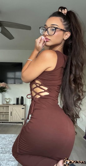 Woman in brown lace-up dress, long dark hair, glasses, posing indoors.