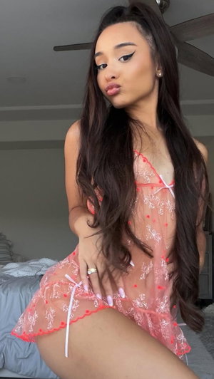 Woman with long dark hair in sheer pink lingerie on bed.
