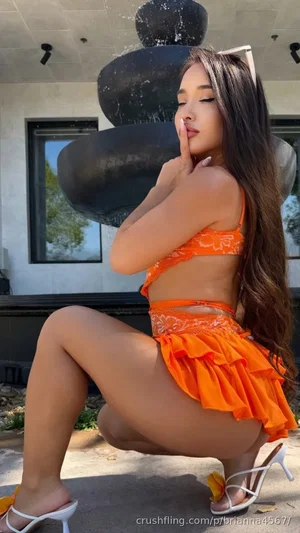 Woman in orange bikini and ruffled skirt posing outdoors near water feature.