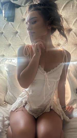 Woman in white lace lingerie sitting on bed with tufted headboard.