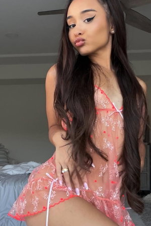 Woman with long dark hair wearing sheer pink lingerie in a bedroom.