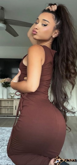 Woman in brown dress posing indoors, long dark hair, makeup, ceiling fan in background.