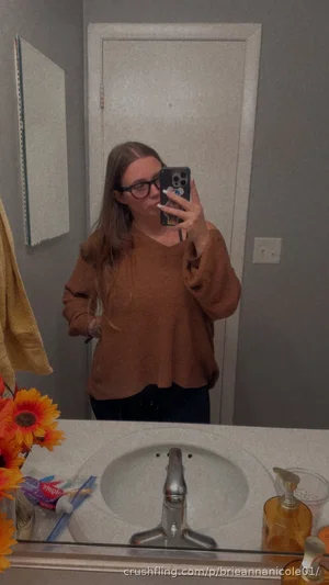 Woman with glasses in brown sweater taking mirror selfie in bathroom.