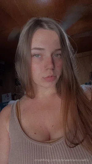 Woman with long brown hair in a beige tank top, indoors, facing camera.