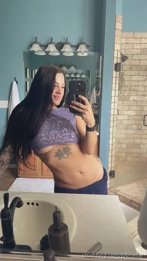 Woman with long dark hair in purple crop top and dark pants, taking mirror selfie in bathroom.