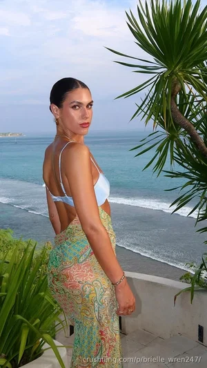 Woman in light blue bikini top and colorful skirt posing by the ocean with palm plants.
