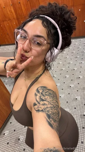 Woman with curly hair, glasses, and tattoos wears brown sports bra and leggings in tiled room.