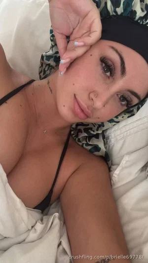 Woman with dark hair and tattoos lying in bed, wearing black bra and headband.