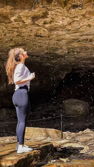 Woman in white top and dark leggings standing under rocky overhang, water dripping around her.