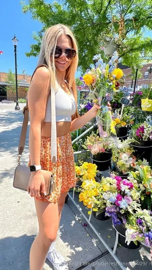 Woman with blonde hair in white crop top and floral shorts holding flowers outdoors.
