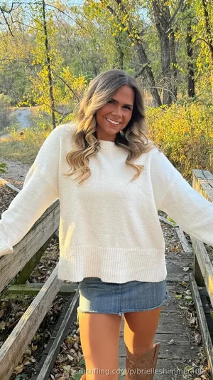Woman in white sweater and denim skirt standing on wooden bridge outdoors.