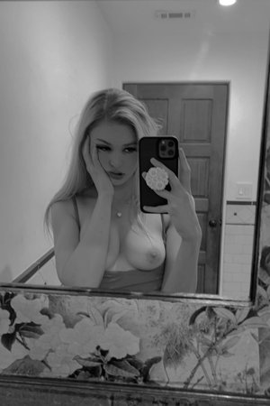 Blonde woman in a low-cut top taking mirror selfie in a room with floral decor.
