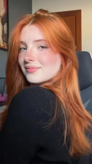 Woman with long red hair wearing black top, sitting indoors in front of chair.