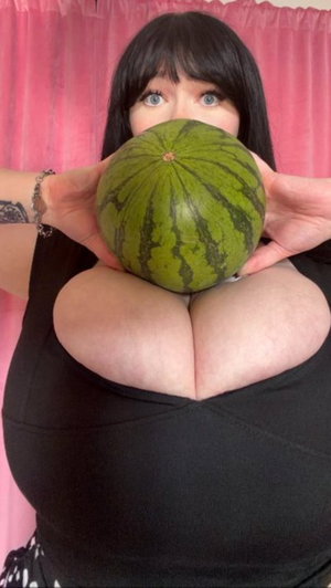 Woman with black hair holding watermelon over chest, wearing black top, pink curtain background.
