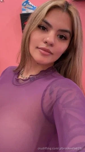 Woman with long blonde hair wearing sheer purple top, posing indoors against pink wall.