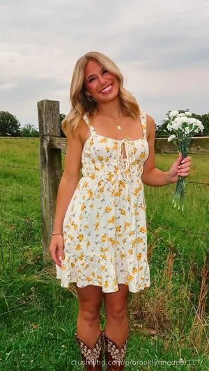 Woman in floral dress and cowboy boots holding flowers in a grassy field.
