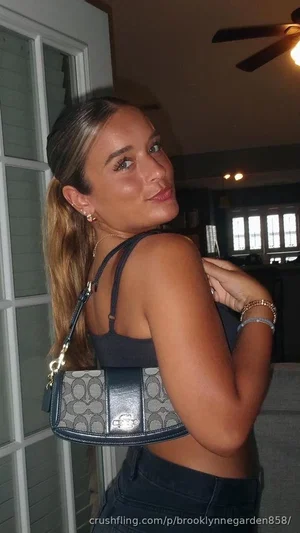Woman with long hair in ponytail wearing black crop top and jeans, holding handbag indoors.