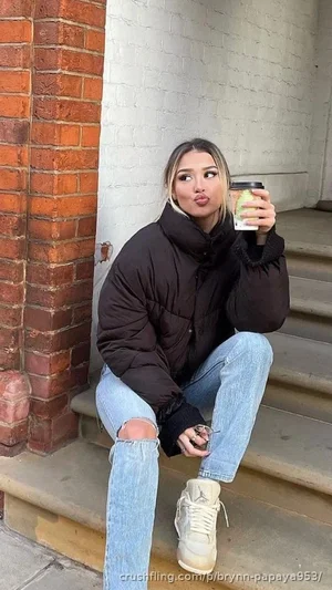 Woman in black puffer jacket and ripped jeans sits on steps holding coffee cup.