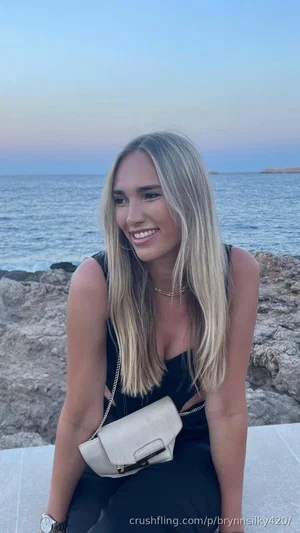 Woman with blonde hair in black dress sitting by ocean rocks, smiling.