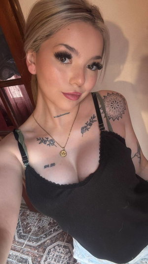 Woman with blonde hair and tattoos wears black top, takes mirror selfie indoors.
