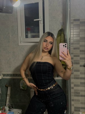 Blonde woman taking mirror selfie in bathroom wearing denim outfit.