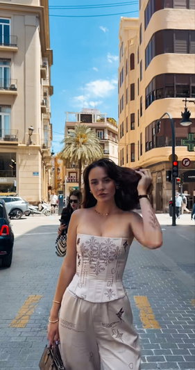 Woman in beige corset and pants walking on city street, holding hair, tattoos visible.