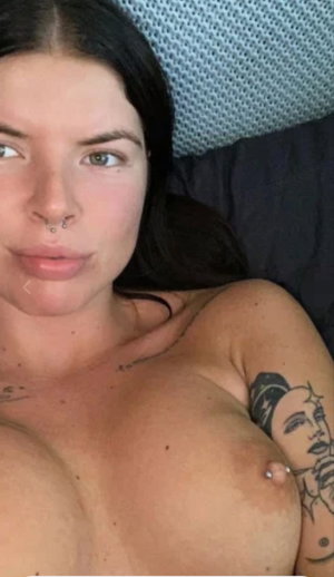 Woman with dark hair and tattoos lying topless in bed.