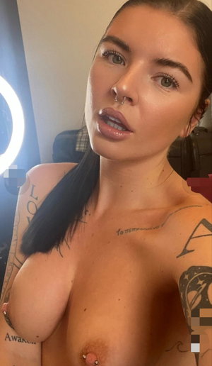Tattooed woman with dark hair taking a topless selfie indoors.