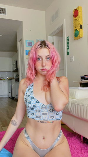Pink-haired woman in patterned crop top and underwear sitting indoors.