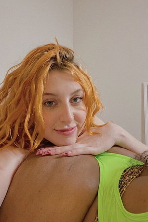 Woman with orange hair in green top resting head on shoulder, indoor setting.