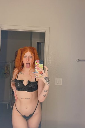 Woman with red hair in pigtails, wearing black cat-themed lingerie, taking mirror selfie in bedroom.