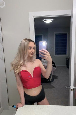 Blonde woman in red top and black shorts takes mirror selfie in bathroom.