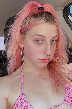 Woman with pink hair and piercings wears pink cherry-patterned top in car.