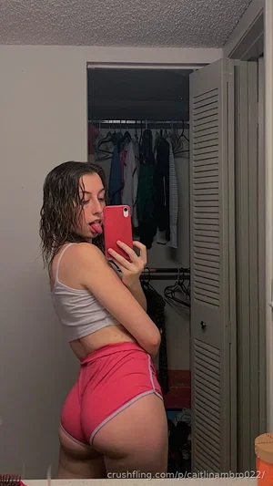 Woman in gray crop top and pink shorts taking mirror selfie in bedroom.