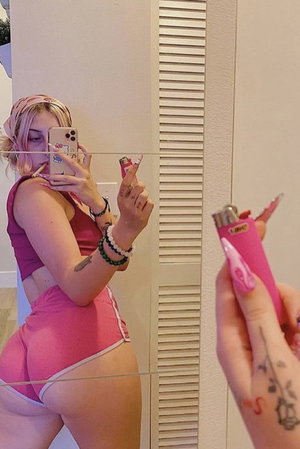 Woman in pink sports bra and shorts takes mirror selfie, holding pink lighter.