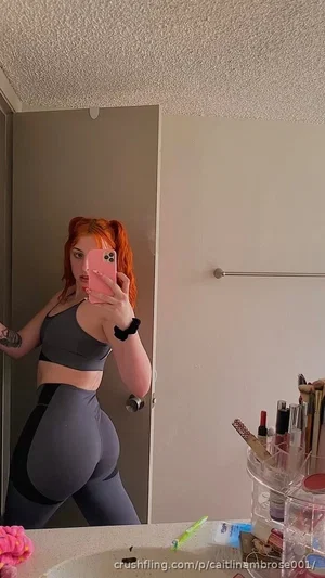 Woman with red hair in pigtails, wearing gray sports bra and leggings, taking mirror selfie in