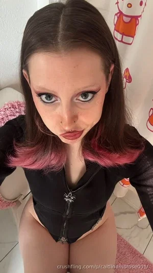 Woman with pink-tipped hair in black zip-up top, posing indoors near Hello Kitty shower curtain.