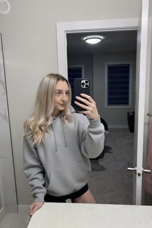 Blonde woman in grey hoodie taking mirror selfie indoors.