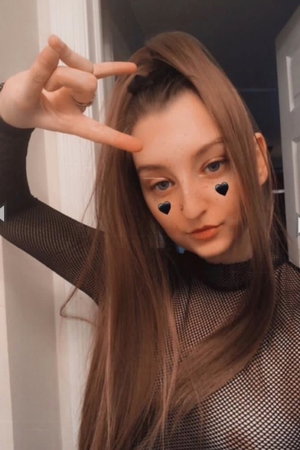 Woman with long brown hair in mesh top making peace sign indoors.