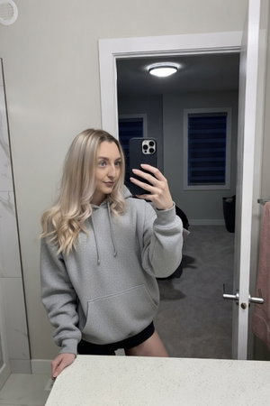 Blonde woman in grey hoodie taking mirror selfie indoors.