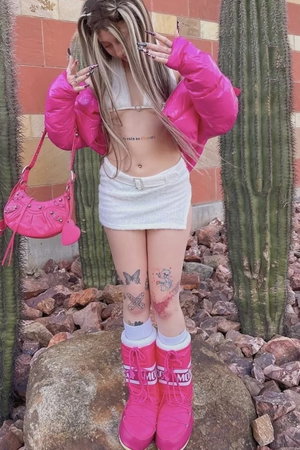 Blonde woman in pink jacket and white skirt outdoors near cacti.