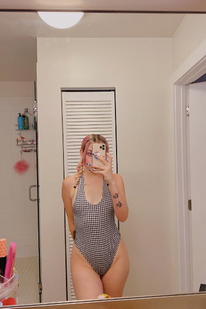Woman with pink hair in checkered swimsuit taking mirror selfie in bathroom.