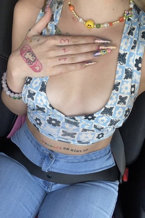 Woman in floral crop top and jeans, hand over chest, tattoos and colorful nails visible.