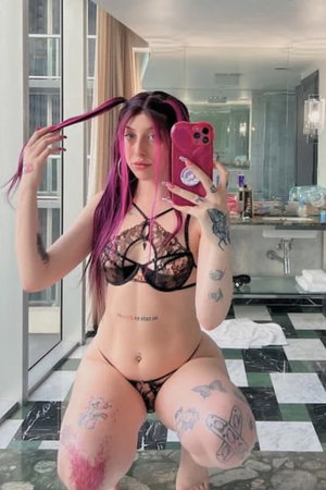 Woman with pink hair and tattoos in black lace lingerie, kneeling in a bathroom.