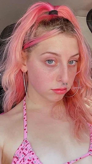 Woman with pink hair and blue eyes wearing a pink cherry-patterned top, sitting indoors.