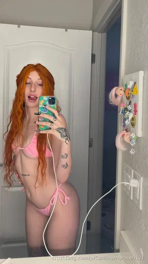 Woman with red hair in pink bikini taking mirror selfie in bathroom.