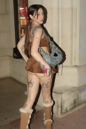 Woman with long brown hair in brown outfit and boots outdoors.