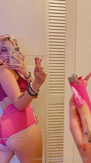 Woman in pink lingerie taking mirror selfie, holding pink lighter, tattoos visible.