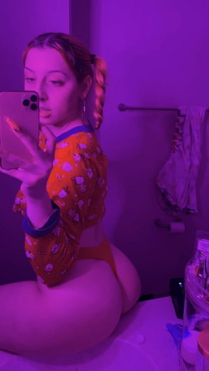 Woman in orange patterned top and underwear, sitting on bathroom counter, taking mirror selfie